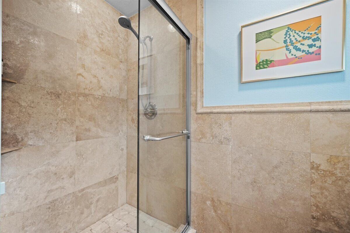 A glass-enclosed shower with a handrail, beige marble tiles, and a framed artwork on the wall.