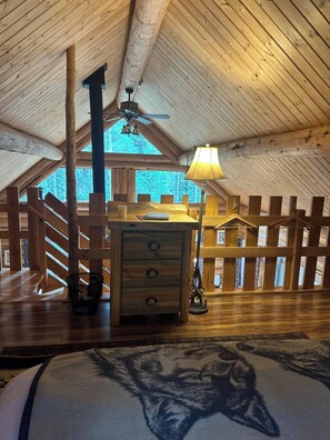 A spacious attic room with wooden beams, a ceiling fan, a central wooden dresser, and a large window offering a view of the forest. - Lamar Valley Mountain Cabin (Silver Gate)