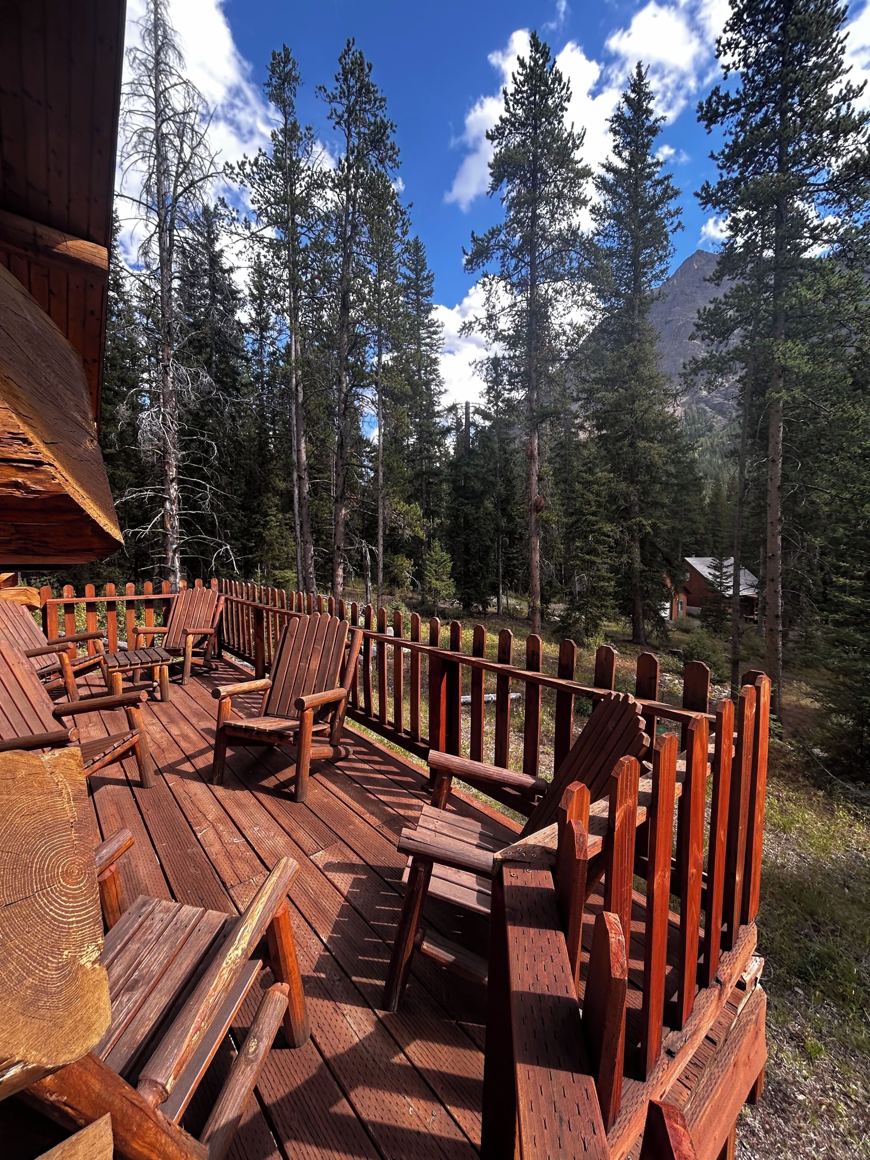 A wooden deck with Adirondack chairs, surrounded by a wooden railing, overlooking a forested mountain area.