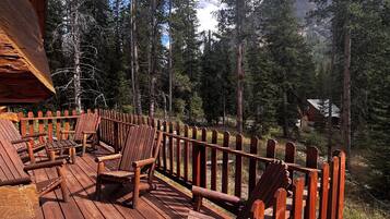 A wooden deck with Adirondack chairs, surrounded by a wooden railing, overlooking a forested mountain area.