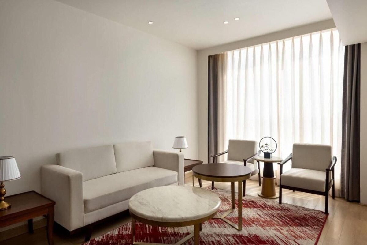 Ein modernes Hotelzimmer mit einer Couch, einem runden Tisch, zwei Sesseln, einer Lampe und Blick auf die Stadt durch große Fenster.