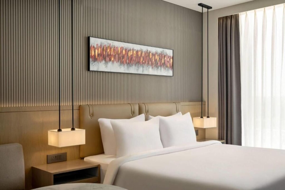 Ein Hotelzimmer mit Bett, Nachttischlampen, einem Kopfteil, einem Wandbild und einem Fenster mit Vorhängen.