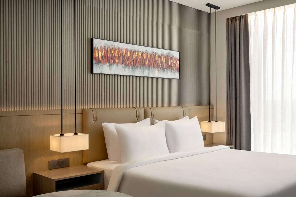 A modern hotel room with a large bed, two bedside tables, a wall-mounted artwork, and a window with curtains.