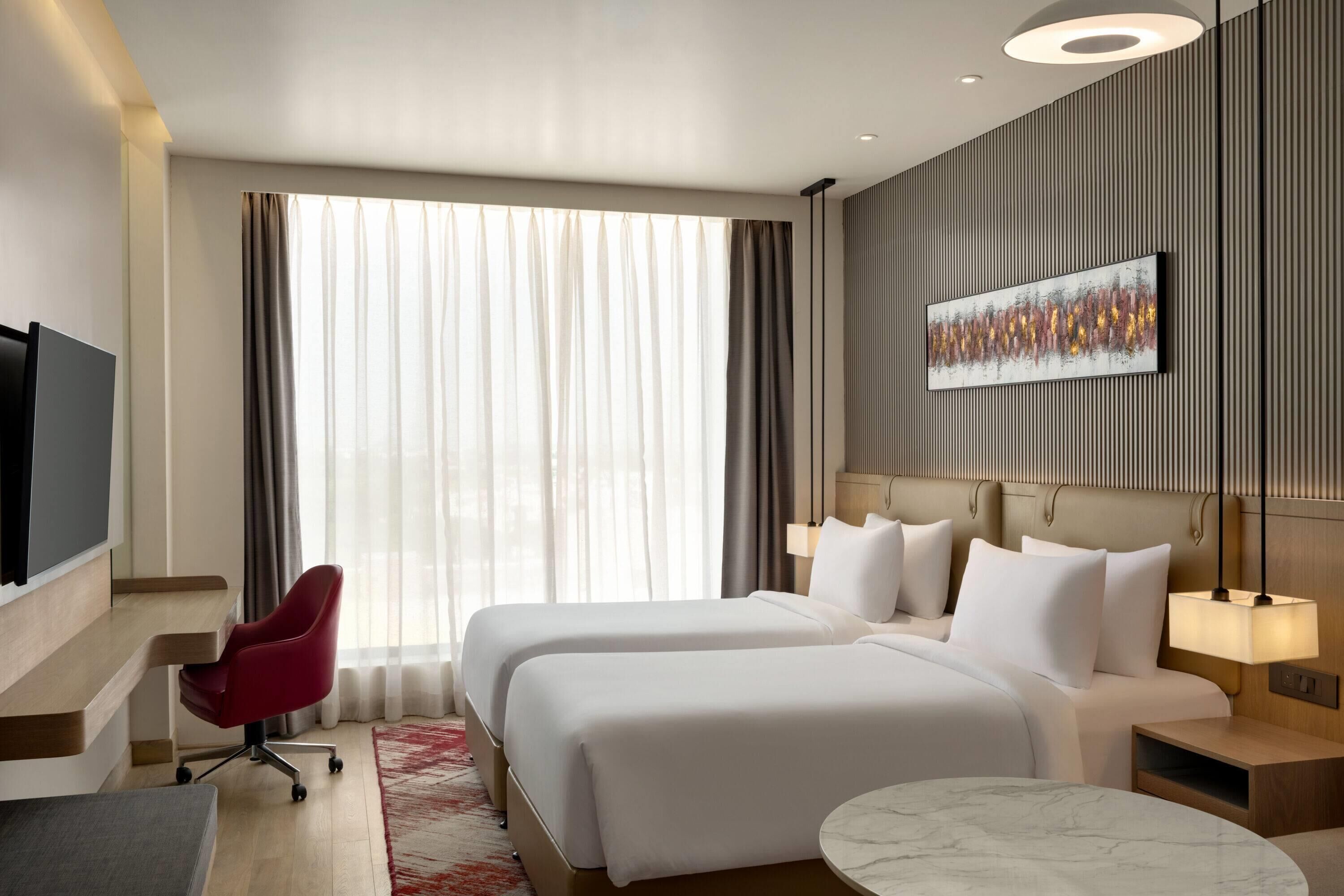 Hotelkamer met twee bedden, een televisie, een bureau, een stoel, een ronde tafel en een groot raam met gordijnen.