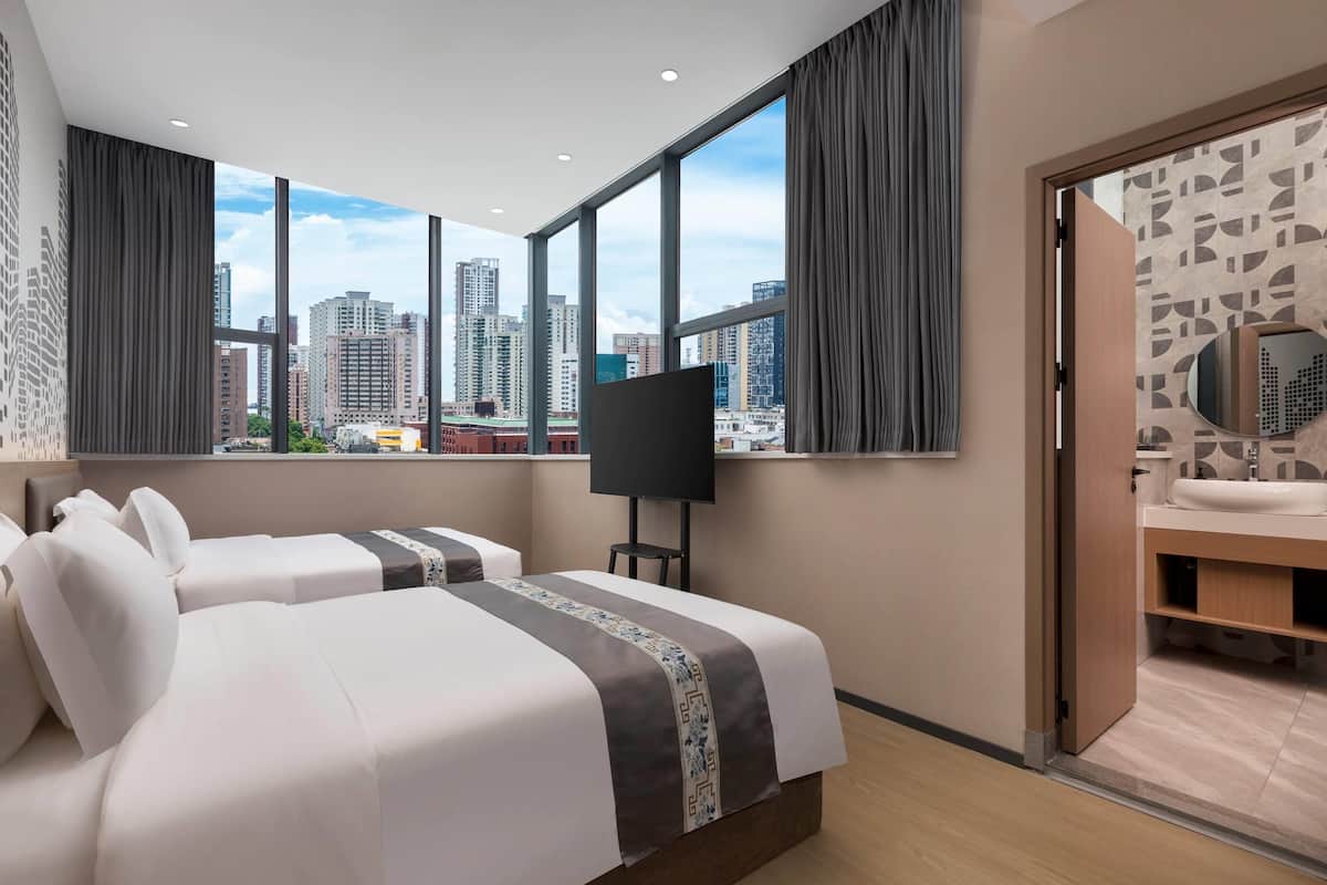 A hotel room with two beds, a large window with a city view, a TV, and a bathroom with a mirror and sink.