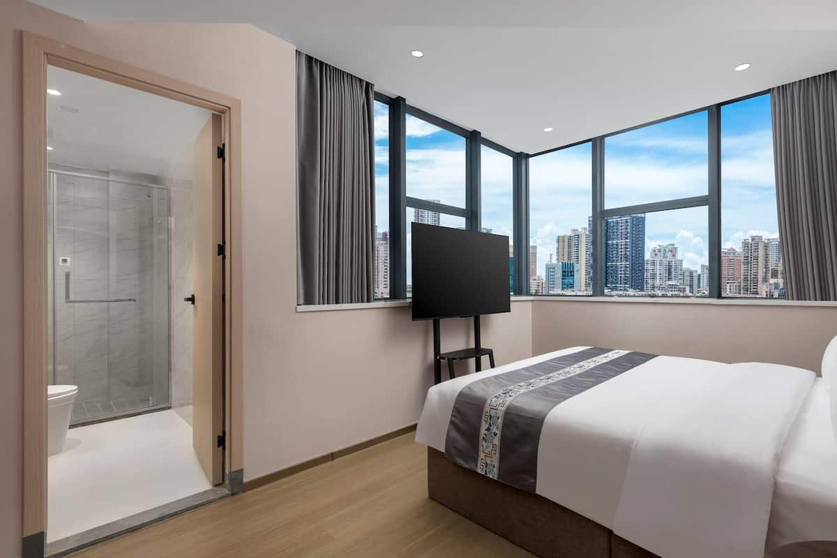 A hotel room with a large bed, a TV, and a city view through large windows.