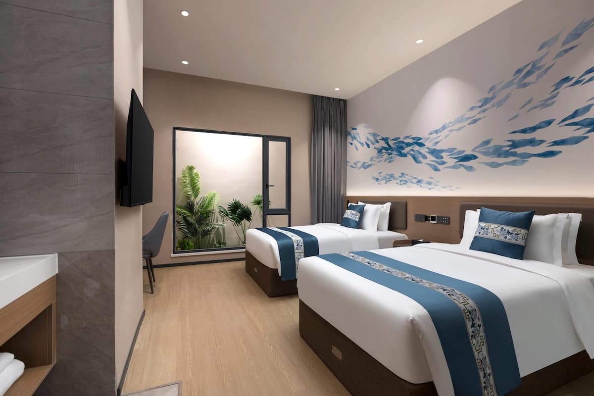 A modern hotel room with two beds, a flat-screen TV, a large window with a view of greenery, and a decorative wall mural.