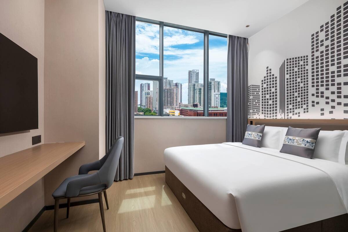 A modern hotel room with a large bed, a desk, and a city view through the window.