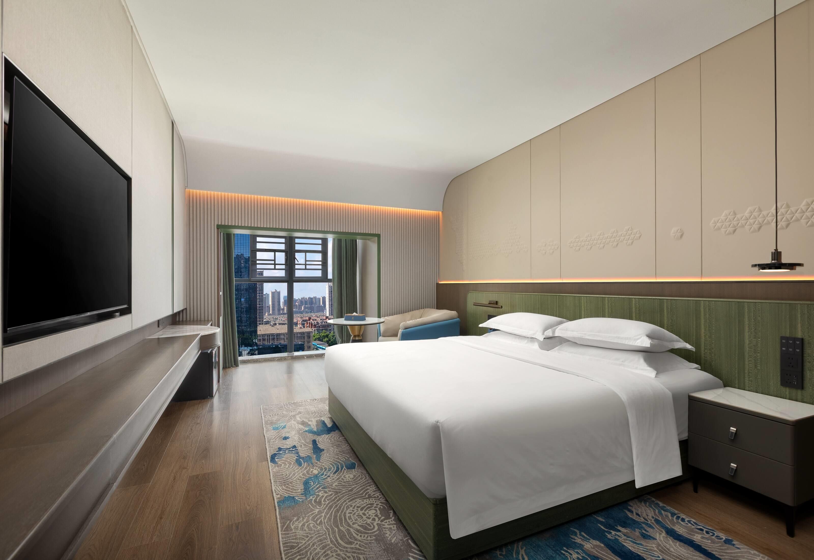 A modern hotel room with a large bed, a flat-screen TV, a view of the city, and a green accent wall.