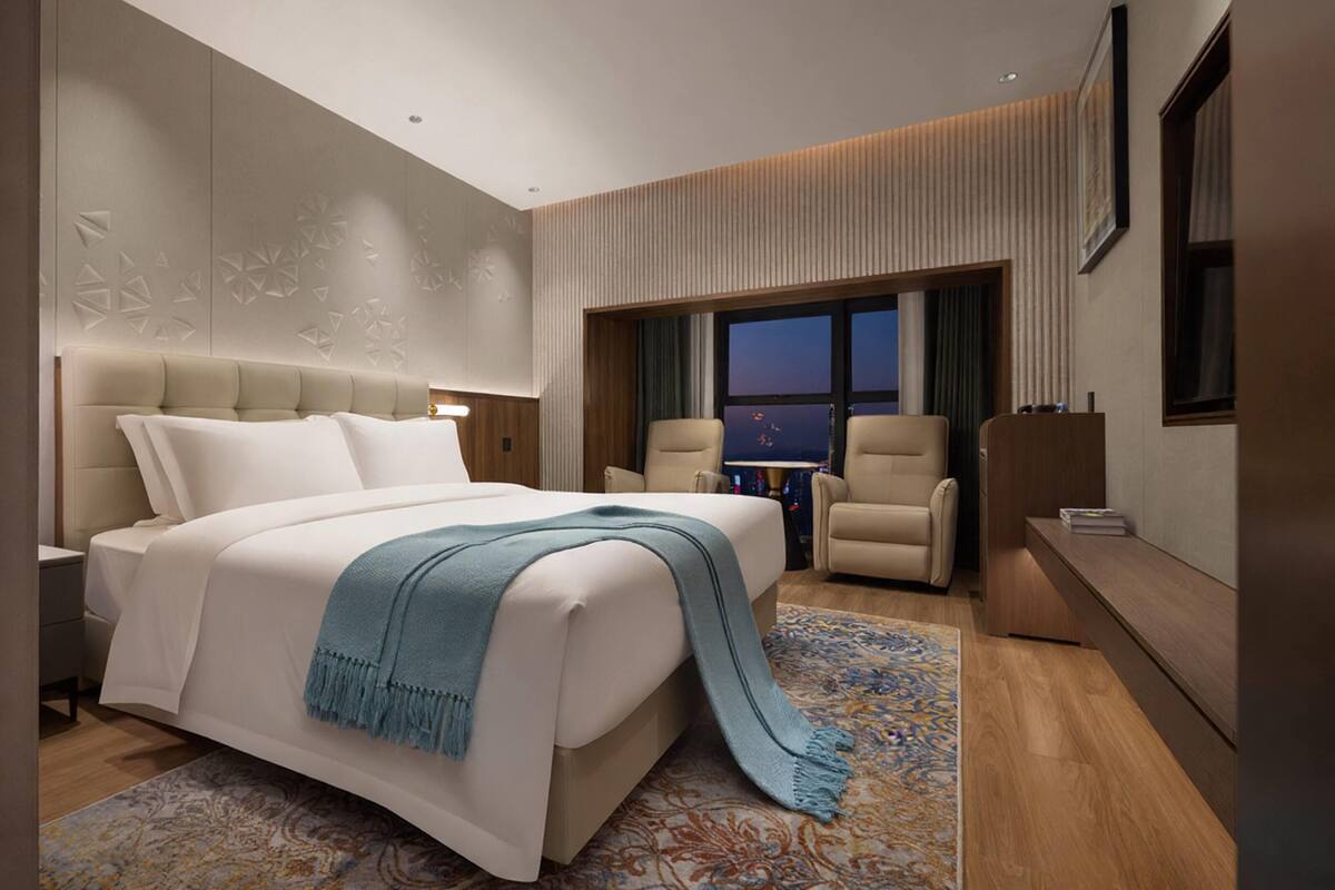Moderni hotellihuone, jossa on suuri sänky, työpöytä, kaksi nojatuolia ja näkymä kaupungin siluettiin auringonlaskun aikaan.