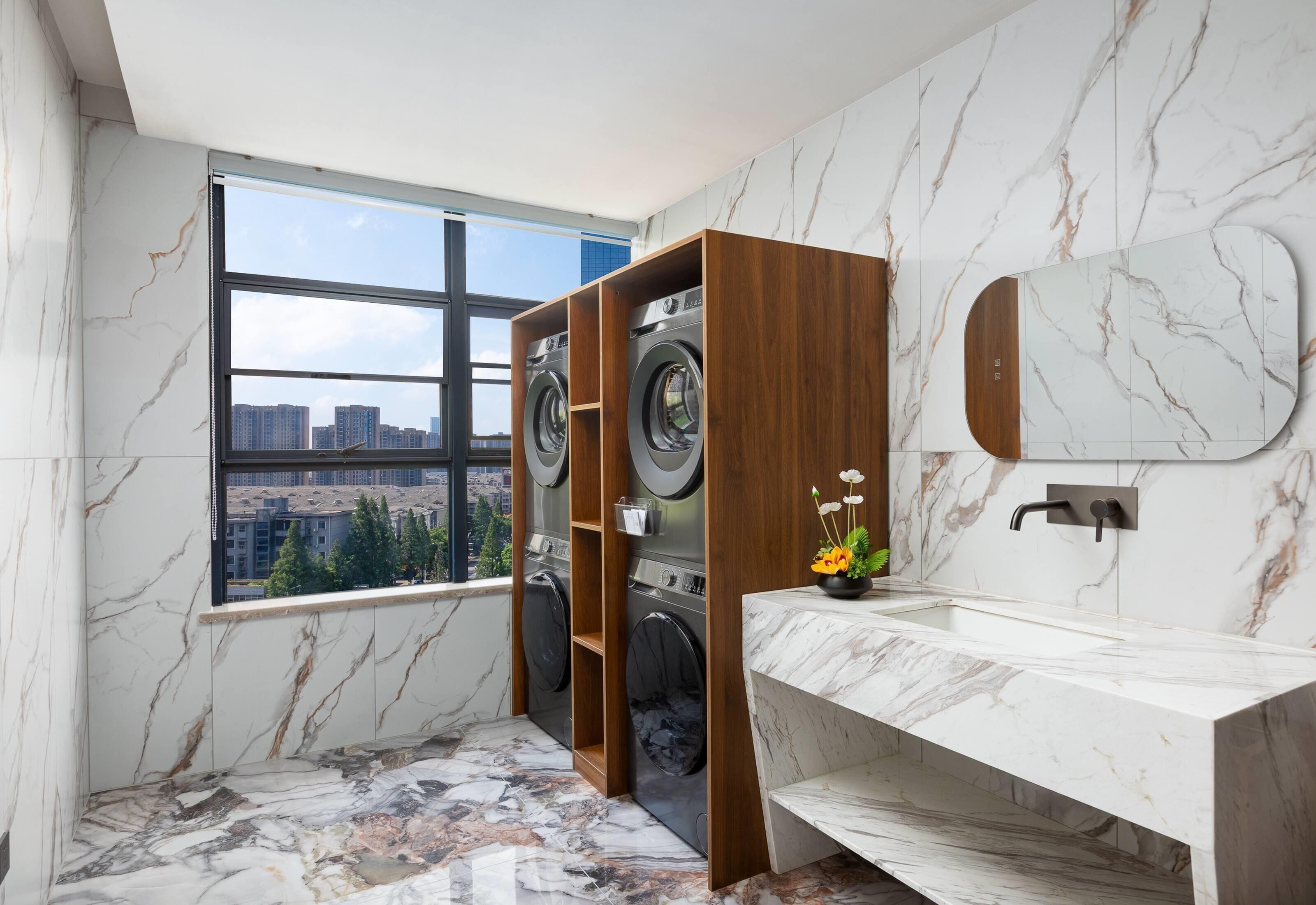 A modern bathroom with marble walls, a large window, a wooden washing station, and a marble sink.