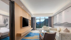 A modern hotel room with a large bed, a TV mounted on the wall, a sofa, a coffee table, and a view of mountains through the window. - Ramada Encore By Wyndham Enshi Huaxi Valley (Enshi)