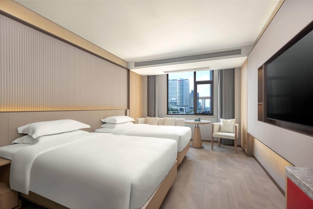A modern hotel room with two beds, a small round table, a chair, and a large TV.