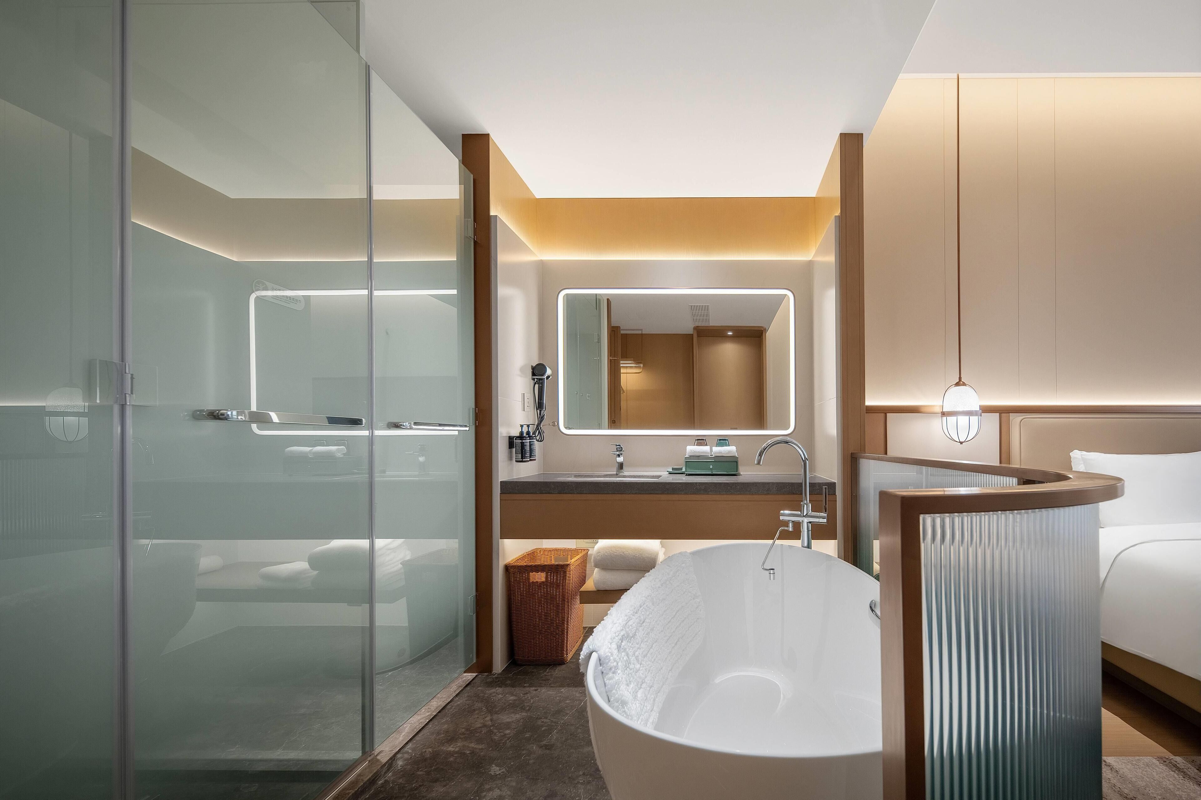 A modern bathroom with a glass shower enclosure, a freestanding bathtub, a vanity with a mirror, and a bedside table.