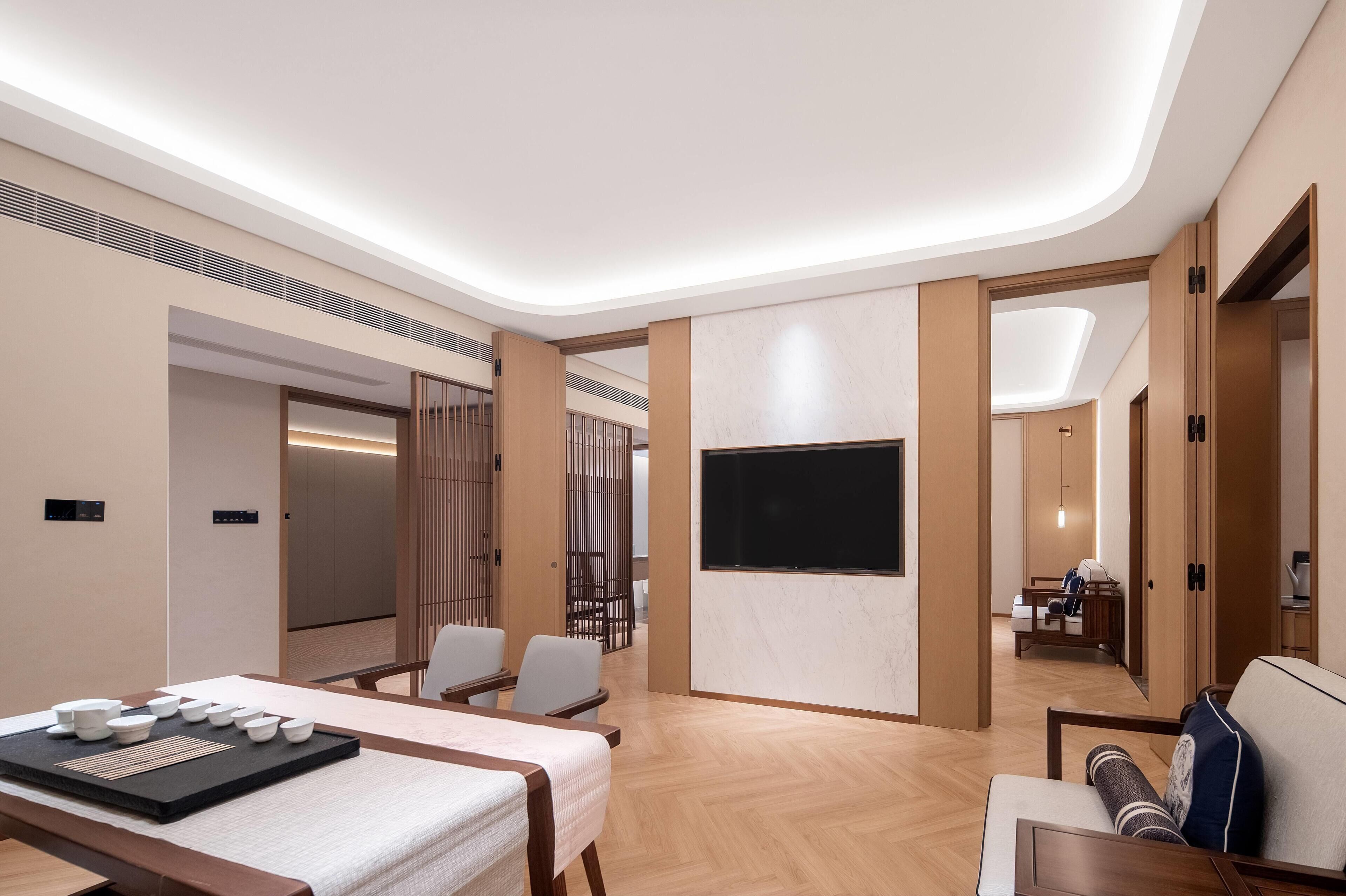 Una habitación de hotel moderna con piso de madera, una cama con cubrecama blanco, un área de descanso con sillas, un televisor de pantalla plana montado en la pared y un panel de madera.