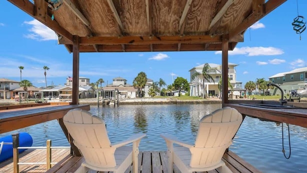 A wooden deck with two white Adirondack chairs facing a calm water body, with a view of residential buildings in the background. - The Gulf Breeze - Direct Access To Gulf (Hernando Beach)
