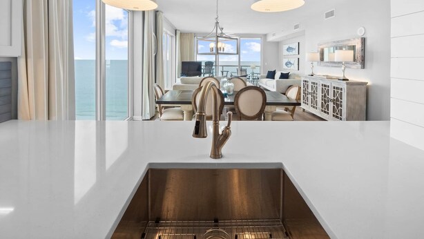 A modern kitchen with a stainless steel sink, a dining area with a table and chairs, and a view of the ocean.