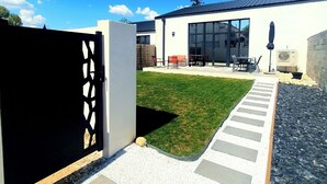 Una casa moderna con un camino de entrada pavimentado que lleva a una puerta de vidrio, un jardín y un patio con mobiliario exterior.