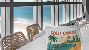 Ein Zimmer mit großen Fenstern mit Blick auf einen Strand, Korbstühle und ein Tisch mit einer 'Cold Coast' Tragetasche.