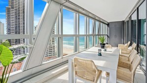 Ein moderner Balkon mit einem weißen Tisch und Korbstühlen, mit Blick auf einen Strand und hohe Gebäude.