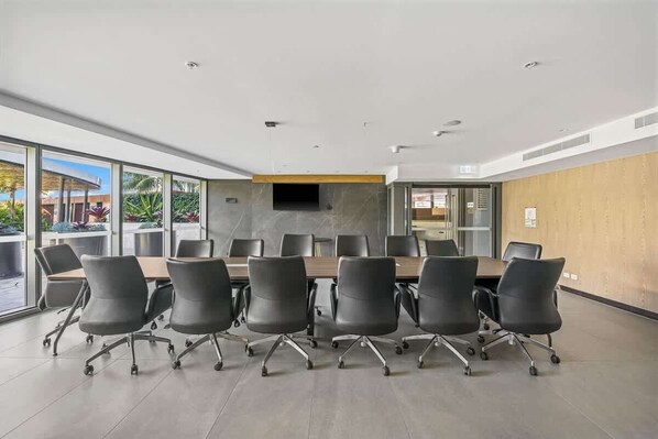 A modern conference room with a long table and black chairs, large windows, and a flat-screen TV on the wall.