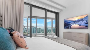 Ein modernes Schlafzimmer mit einem großen Bett, einem an der Wand montierten Fernseher und Blick auf eine Stadtkulisse durch große Fenster.