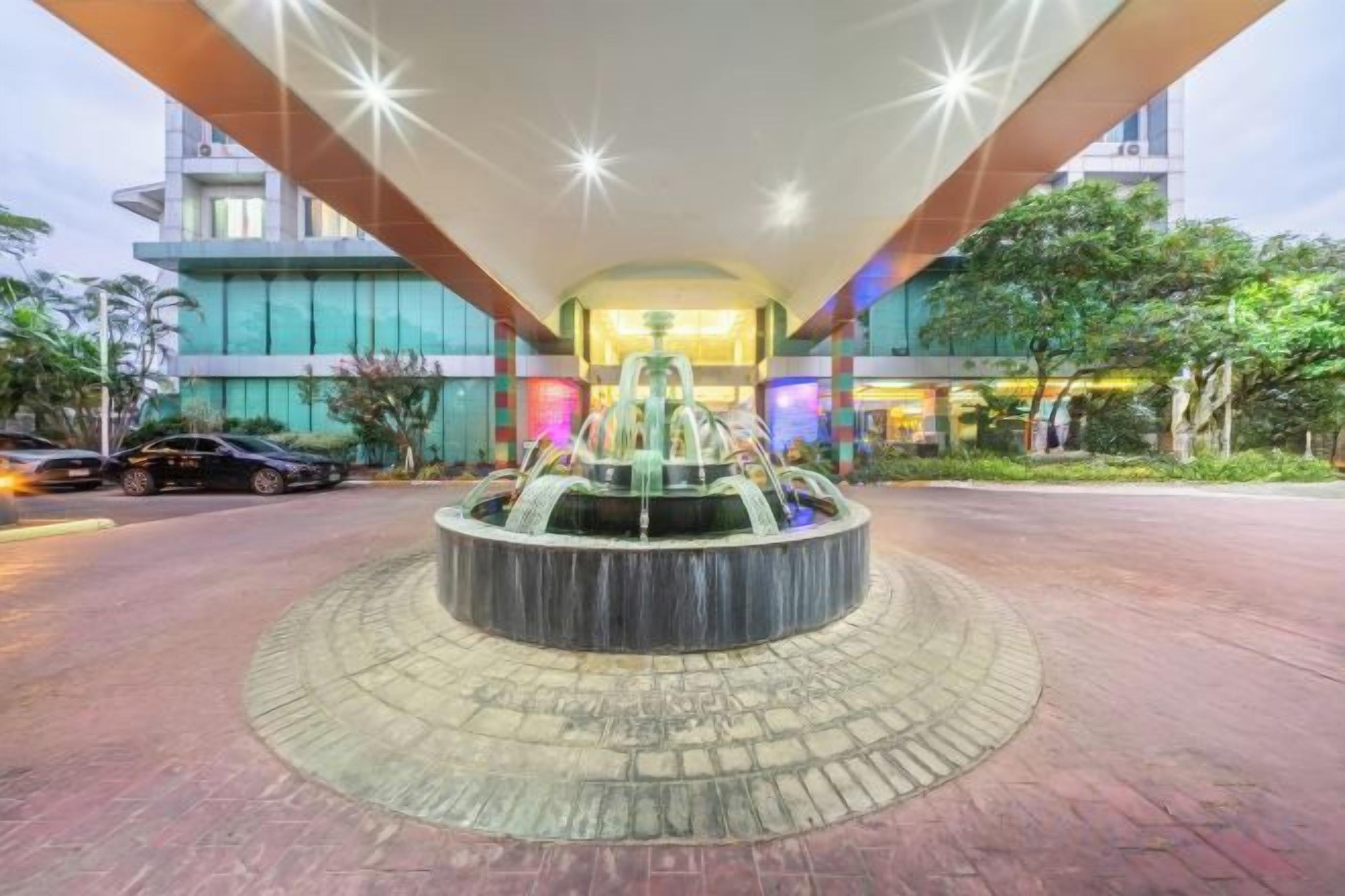 A hotel entrance with a central fountain, paved walkway, and a building with a modern design.