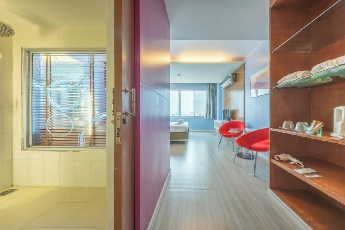 A hotel room with a wooden floor, a red chair, a wooden shelf with items, and a view through a window with blinds.
