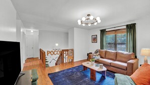 Un salon moderne avec un canapé en cuir, une table basse et un téléviseur. Un escalier mène à une autre pièce, et un mur porte l’inscription « POCO ».