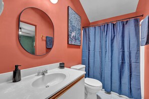 Ein Badezimmer mit einem weißen Waschbecken, einer Toilette, einem blauen Duschvorhang und einem Spiegel mit rundem Rahmen.