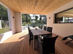 Eine Terrasse im Freien mit einem Holztisch und Stühlen, einem großen Fenster und Blick auf einen Garten und Bäume.