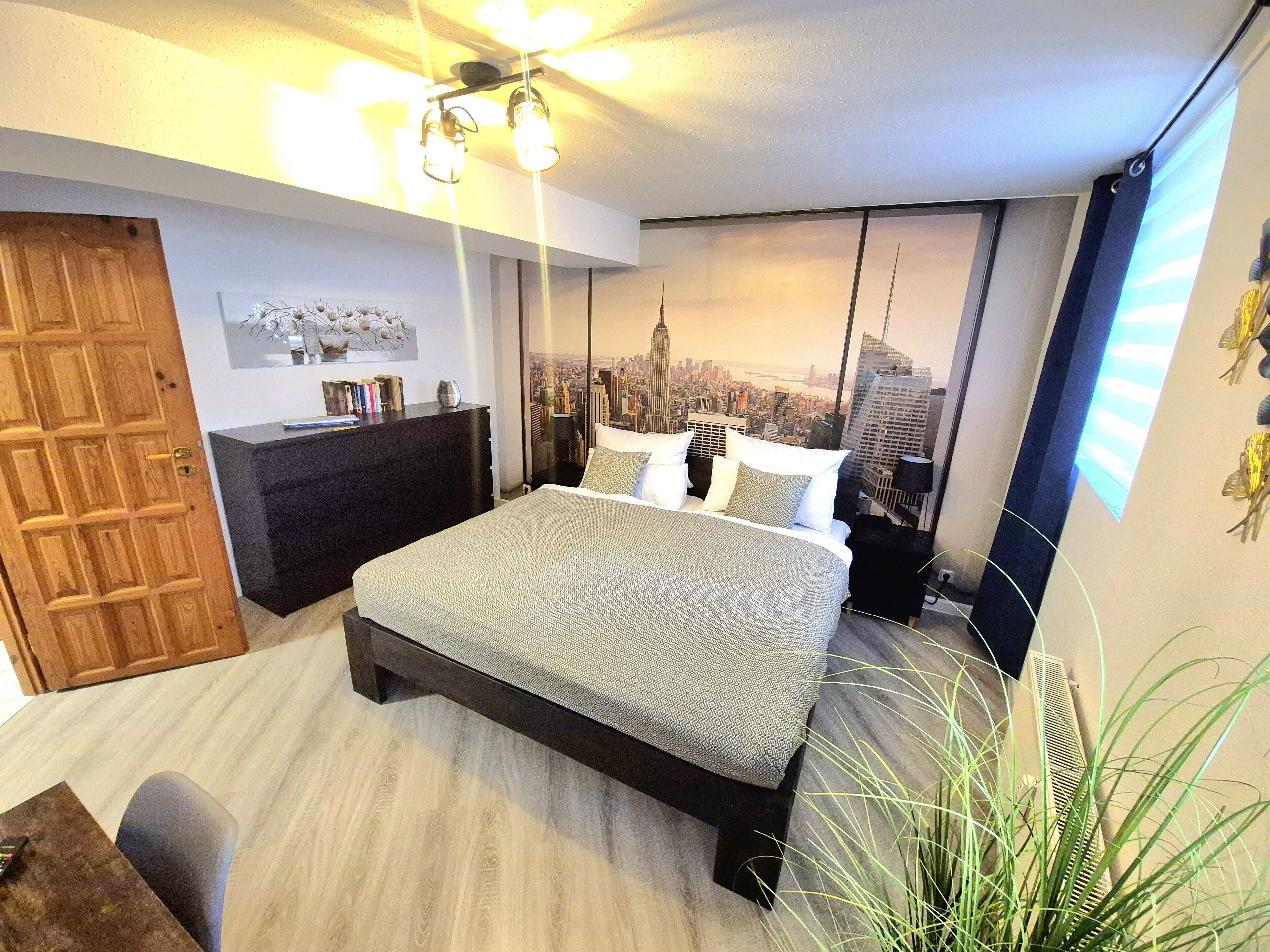 A modern bedroom with a cityscape view, a large bed, a dresser, and a dining area.