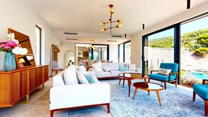 A modern living room with a sofa, coffee table, and chairs, a wooden cabinet with a vase of flowers, and a large window offering a view of greenery. - Villa Flora Mare IV with Two Pools (Primošten)