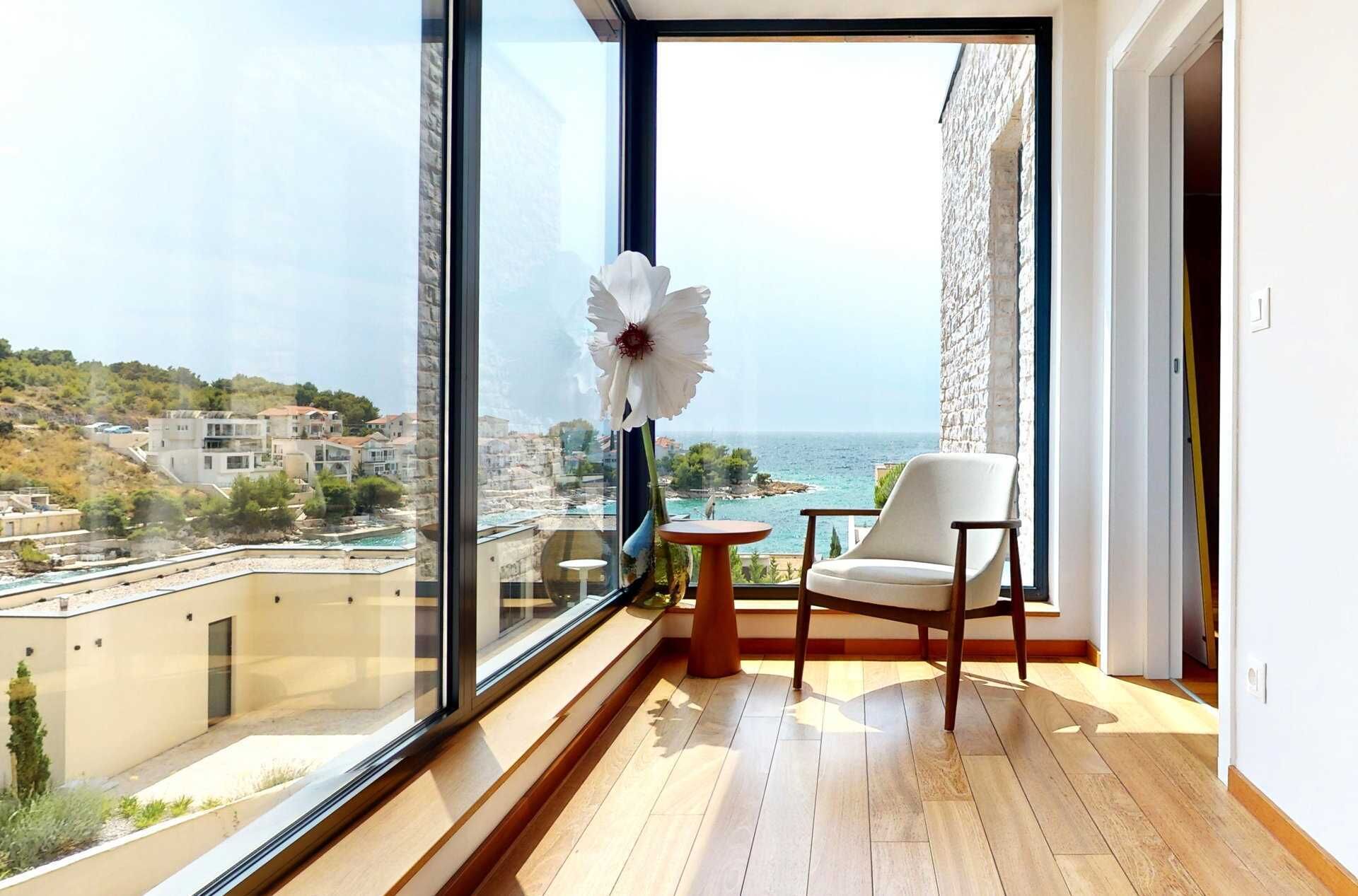 A balcony with a wooden floor, a white chair, and a vase with a white flower. A large window offers a view of a coastal town and the sea.
