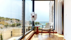 Un balcón con piso de madera, una silla blanca y un jarrón con una flor blanca. Un ventanal grande ofrece vistas a un pueblo costero y al mar.