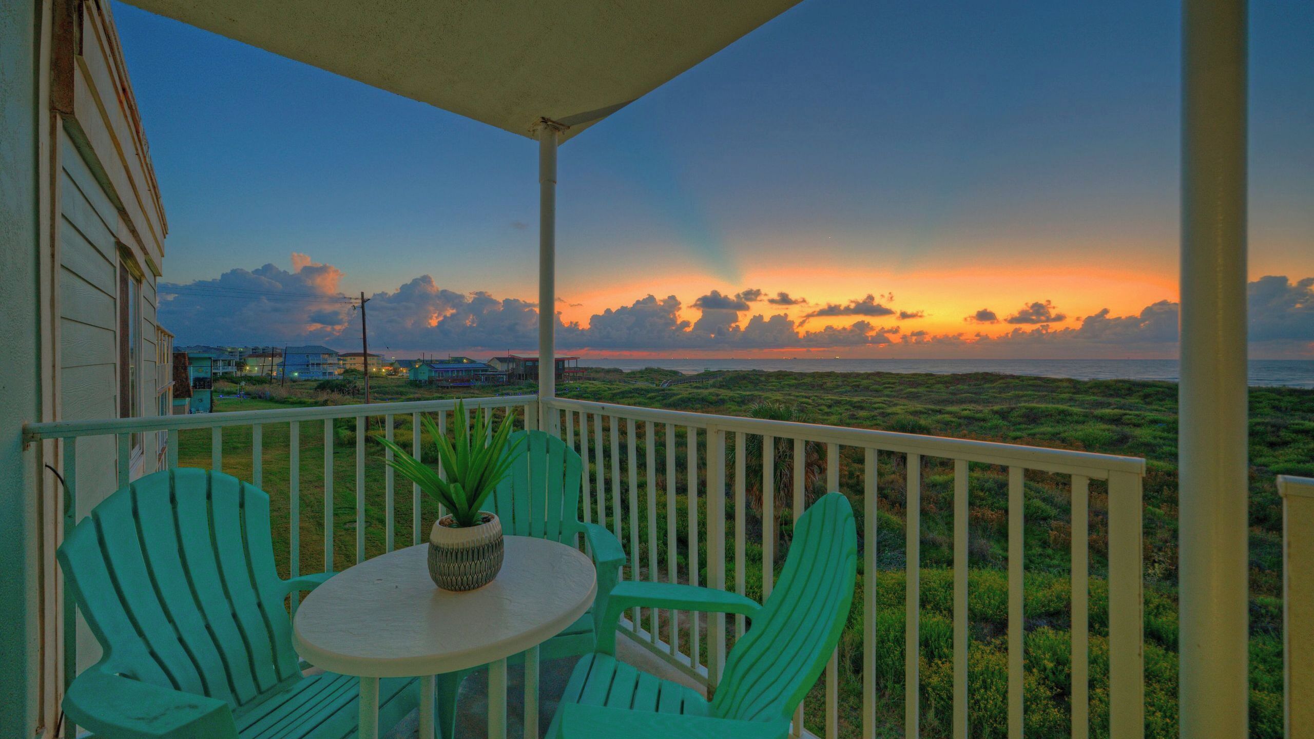 A balcony with a view of the sunset over the ocean, green chairs, and a small table with a plant.
