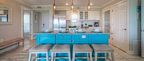 A kitchen with a blue island, four barstools, pendant lights, and a refrigerator.