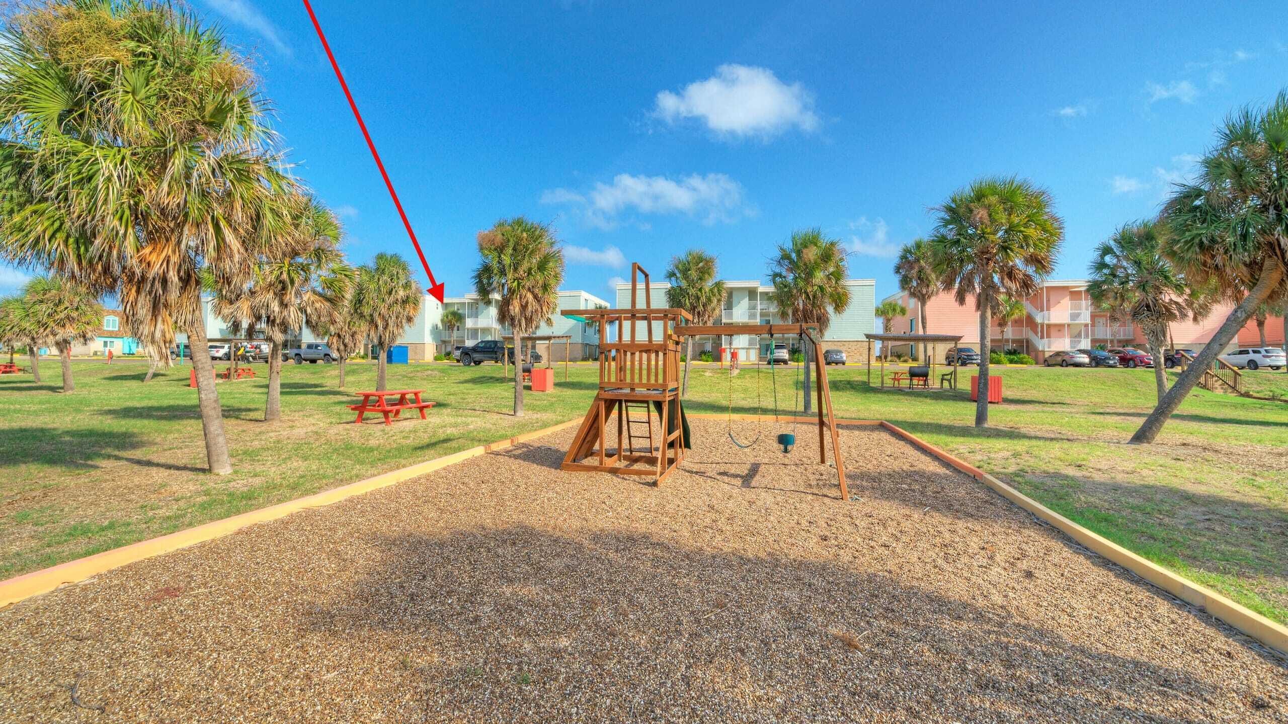 A playground with a wooden swing set and a red bench, surrounded by palm trees and a clear sky.