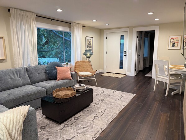 A modern living room with a grey sofa, a black coffee table, and a wicker chair. There is a dining table with white chairs and a lamp. The room has dark wooden flooring and a large window with curtains.