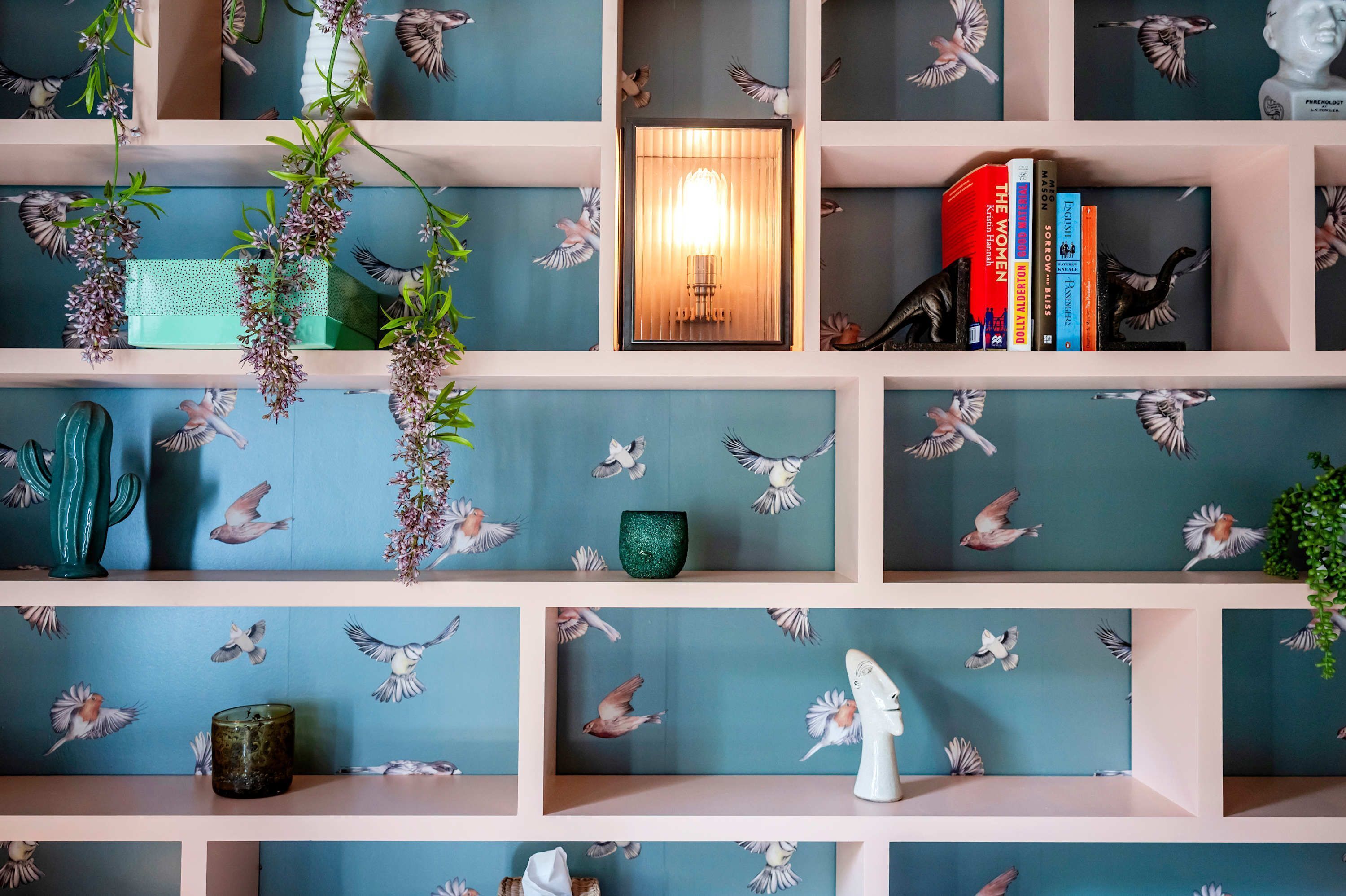Une pièce avec un mur bleu orné d’un motif d’oiseaux, une bibliothèque remplie de livres, un vase vert et une plante suspendue.