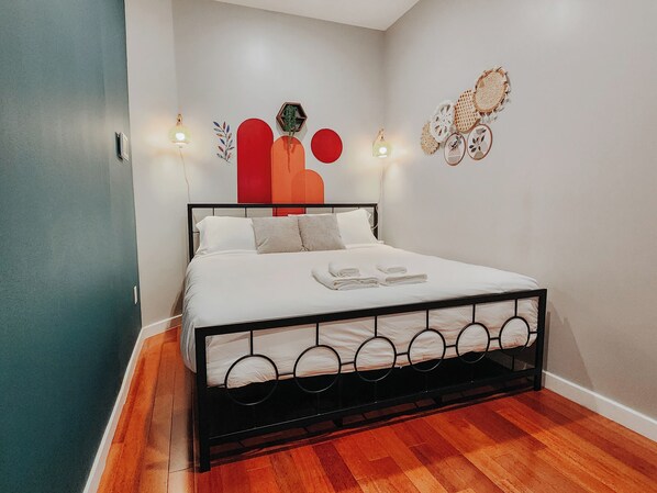 Una cama bien hecha con sábanas blancas y estructura de metal negro, ubicada en una habitación con piso de madera y paredes de color claro.