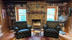 A cozy room with a stone fireplace, two leather armchairs, a large rug, and bookshelves filled with books. - Charming Shared Cabin at Project Chimps Sanctuary (Morganton)