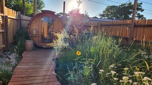 Una cabaña de madera con un porche de madera que lleva a una puerta, rodeada de diversas plantas y pastos.