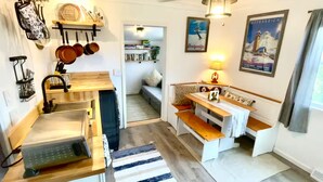 Cocina compacta con lavamanos, estufa y utensilios colgantes. Una zona de comedor con banco y mesa está contigua, con una lámpara y un cartel.