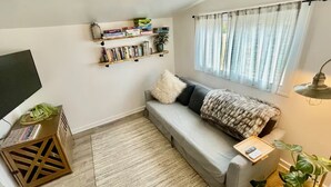 Una sala de estar acogedora con un sofá gris, una estantería, un televisor y una ventana con cortinas translúcidas.