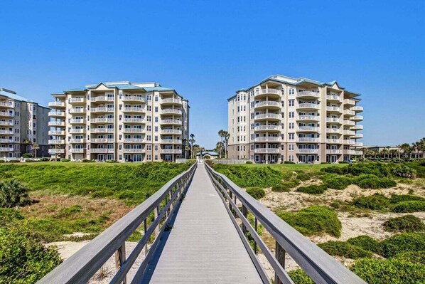 Une promenade en bois menant à des bâtiments situés en bord de mer, dotés de balcons, sous un ciel dégagé, avec une végétation verdoyante.