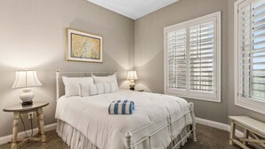 A bedroom with a bed, two bedside tables, a ceiling fan, a window with shutters, and a framed picture on the wall.