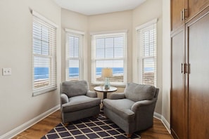 A corner room with three windows, a small round table with a lamp, two grey armchairs, and a wooden wardrobe.