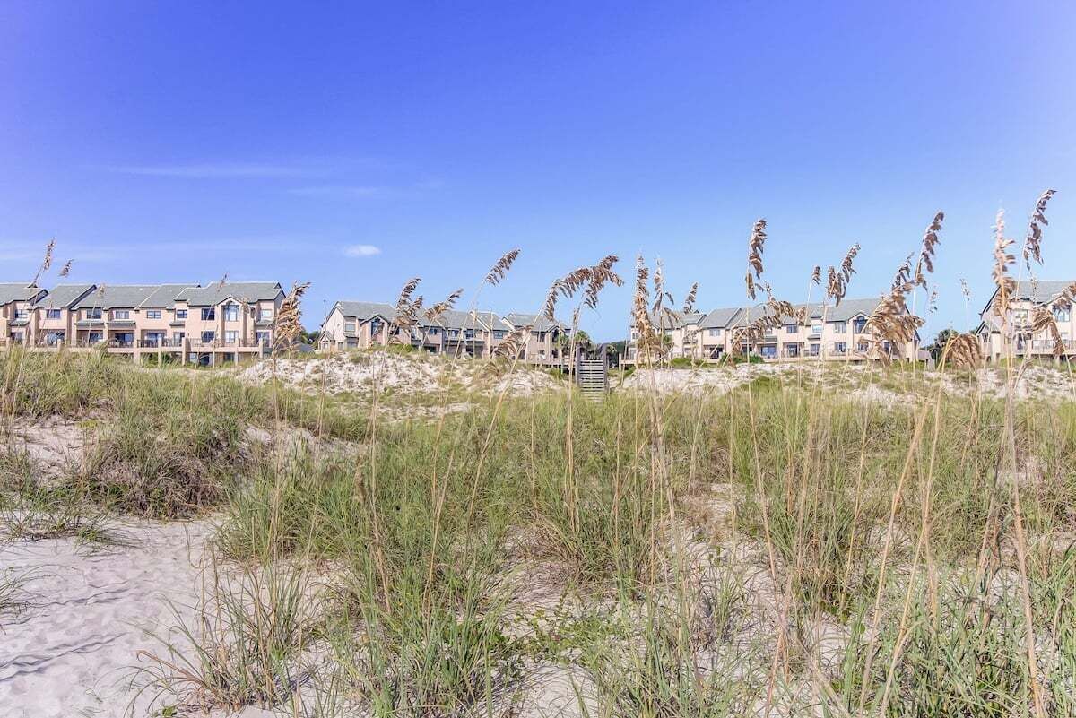 A beachfront hotel with multiple-story buildings, sandy dunes, and tall grasses.