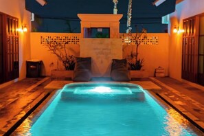 Une piscine extérieure avec un mur en pierre, deux chaises et une petite fontaine.